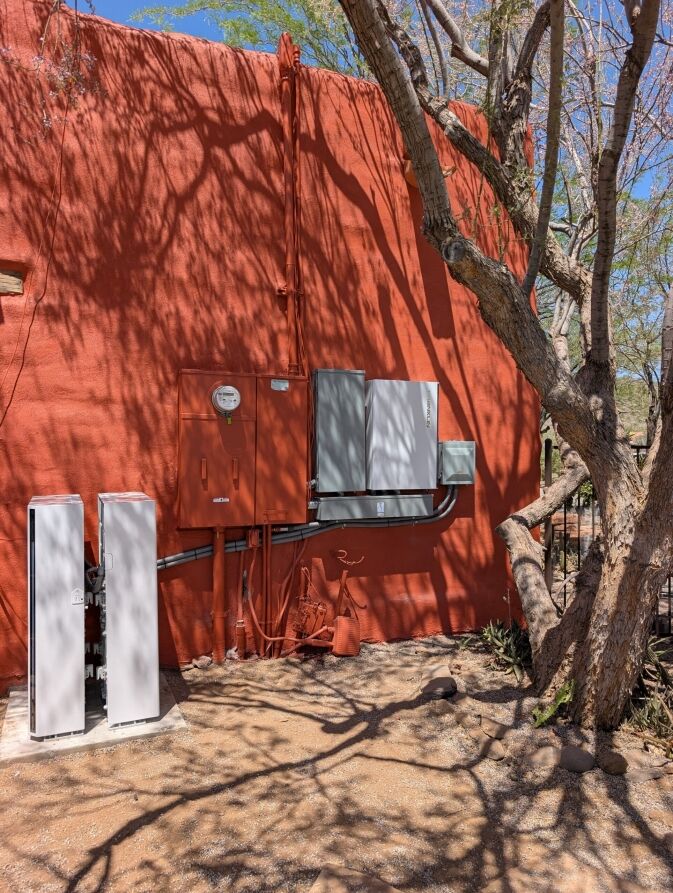 Electrical equipment, including meters and wall-mounted boxes for electrical service upgrades, installed on a bright orange exterior wall next to a tree.